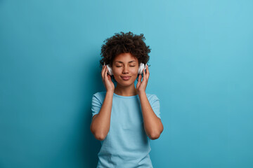 Satisfied curly young Afro American woman has carefree happy mood, closes eyes and listens music in headphones, wears casual blue t shirt, poses indoor. People, leisue, entertainment concept