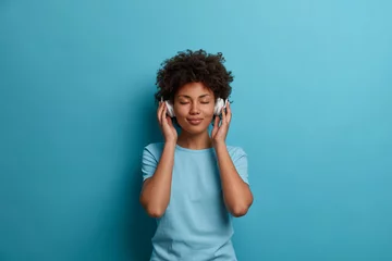 Poster Satisfied curly young Afro American woman has carefree happy mood, closes eyes and listens music in headphones, wears casual blue t shirt, poses indoor. People, leisue, entertainment concept © Wayhome Studio