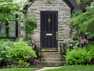 front door of stone faced house with leaded glass windows, surrounded by flowers and shrubs