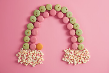 Cretive shot made of colorful french macaroons and popcorn. Rainbow of sweet cookies on rosy background. Pistachio, stawberry, orange flavor. Appetizing dessert. Tasty life and food concept.