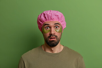 Young stunned unshaven European man stares with disbelief, finds out shocking news, wears bath cap and green undereye patches for reducing wrinkles and puffiness, isolated on green background