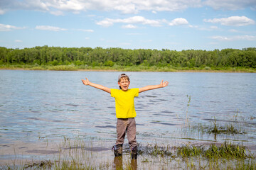 Fototapeta na wymiar happy boy in boots stands in the river, arms spread wide