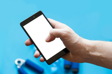 Smartphone with white screen in mans hand and sports equipment on blue background.