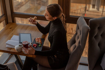 Female posing in cafe  on the roof of the city and using a laptop.  Fashion woman dressed style black  golf and shorts. Freelancer relexing on the rooftop and holding computer, eating sushi.