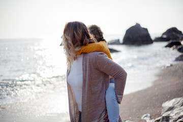 Happy family posing outdoor in the beach of the sea at spring time. woman with daughter have fun on vacation near ocean. Female parent rolls the child on shoulder. 