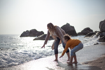Family posing outdoor in the beach of the sea at spring time. woman with daughter have fun on vacation near ocean. Female parent holds childs hand and touch the water. 