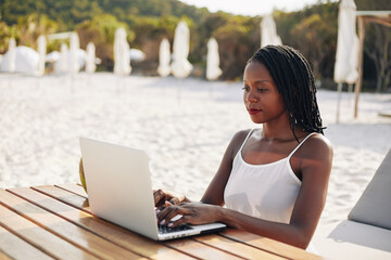 Smiling pretty young travel blogger sitting at wooden table on beach and working on laptop when...
