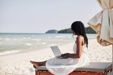 Beautiful young woman with dreadlocks sitting on chaise-lounge, working on laptop and enjoying sound of waves