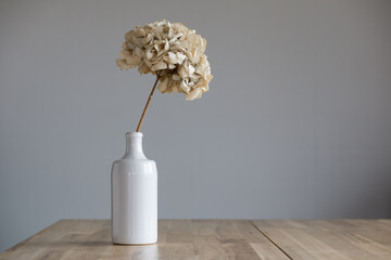 A white ceramic bottle with a dry hydrangea flower stands on a wooden surface.