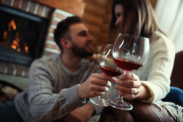 Couple with glasses of wine near fireplace indoors, focus on hands. Winter vacation