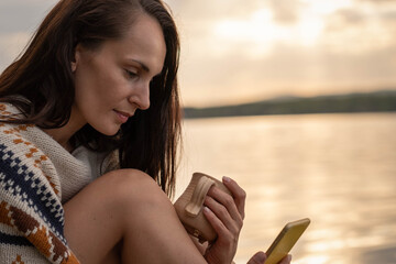 A beautiful dark haired woman sits on an old wooden pier in the early hours of dawn holding a smartphone and a ceramic coffee mug