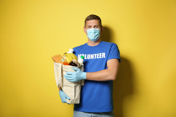 Male volunteer in protective mask and gloves with products on yellow background. Aid during...