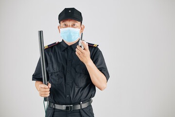 Portrait of security officer in medical mask holding truncheon and talking on walkie-talkie
