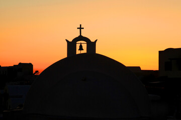 The silhouette of a church with crucifix and bell against an orange sunset sky on the island of Santorini, Greece in the Aegean Sea.