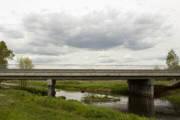 A red passenger car rides on a small concrete bridge over a shallow small river in the summer in the countryside. Above the bridge are large clouds and clouds.