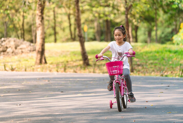 Fototapeta premium Front view Asian child girl practicing biking bicycle with one supporter wheels. Happy with bicycle, smiling face girl, 6 years old girl. Blurred background tree in park, space for text and design.
