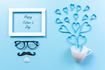 Happy Fathers Day background concept.Decoration light blue heart, frame, mustache, eyeglasses splash out of coffee cup on light blue pastel background with copy space. Top view, flat lay.