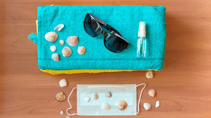 A sanitizer bottle, face mask, sunglasses and shells on a blue towel against a wood background. The concept of a summer vacation in a new reality.