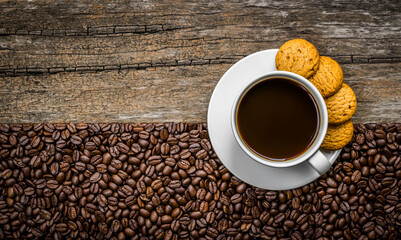 Top view of coffee in white cup with cookie on white saucer and coffee beans on wooden background.