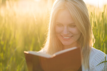 Attractive smiling young woman reading a story book outdoor inside a green meadow field with...