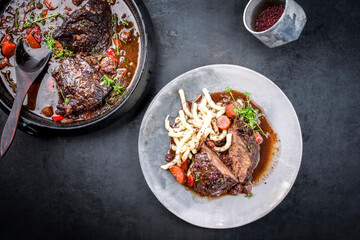 Traditionelle deutsche geschmorte Ochsenbäckchen in brauner Rotwein sauce mit Nudeln und Karotten angeboten als Draufsicht auf einem Modern Design Teller und Schmortopf stewpot auf einem Board