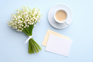 Mothers Day or Wedding flat lay composition with cup of coffee, bouquet of lily of the valley flowers, love letter with blank white note on blue background. Top view.