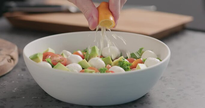 Slow motion man squeeze orange lemon into salad with kale, avocado, mozzarella and tomatoes in white bowl on kitchen countertop