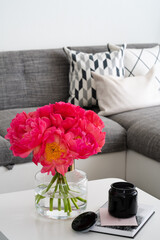 Bouquet of the pink peonies and book with black candle on the white table and sofa with pillows on the background.
