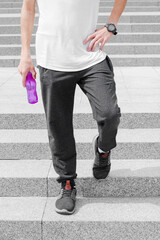 Young sporty man in grey sportswear walking on stairs outdoors with a bottle of water from eco plastic. Concept of sport, outdoors activity, rest, health care, environmental protection.