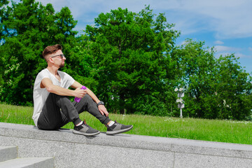 Young sporty man sitting outdoors in park with a bottle of water from eco plastic. Resting man in sunglasses on a green background. Concept of sport, outdoors activity, rest, health care.
