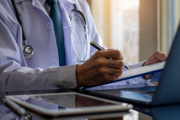 Doctor man in white lab coat writing note on data chart clipboard with laptop computer and digital...