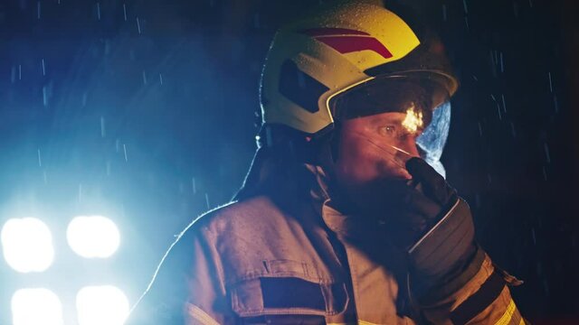 Portrait shot of a fireman in action speaking on the walkie talkie. Fire reflection on the helmet and reflectors in the background. Slow motion.