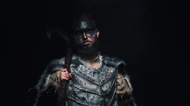 An aggressive Viking in wool armor puts an axe on his shoulder and glares at the camera. A Viking in armor on a black background with a two handed axe looks at the camera