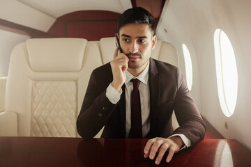 Handsome rich business man wearing suit and tie, sitting in private jet next to window, answering phone call with concentrated face