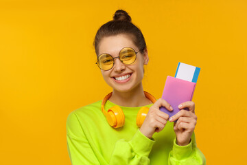 Young happy smiling teenage girl in neon green sweatshirt and glasses holding passport with airplane tickets, ready for vacation, isolated on yellow background