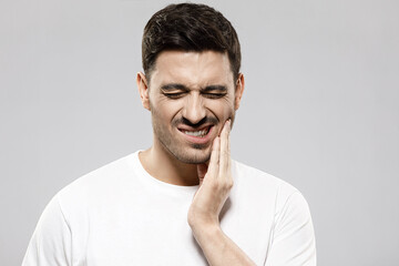 Fototapeta premium Toothache concept. Portrait of young man in white t-shirt holding his cheek trying to ease severe tooth pain, isolated on gray background