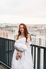 Beautiful girl on the terrace in a white blanket at sunset. Girl posing background fashion. Mood girl wrapping with blanket on balcony. Terrace mood.