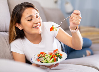 Peruvian housewoman eating vegetable salad on sofa at home