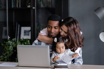 Happy friendly mixed race a family african-american father asian mother and charming daughter are sitting at a table with a laptop and a notebook. Freelance work and happy family concept