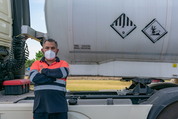 Truck driver with face mask posing next to a dangerous goods tank truck