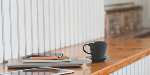 Close up image of the coffee cup is putting on a wooden plank that surrounded by laptop, tablet, smartphone, and equipment.