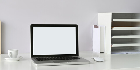 A white blank screen computer laptop is putting on a white workspace surrounded by office equipment.