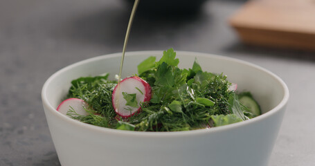 pour olive oil into fresh salad with radish, cucumber and herbs in white bowl