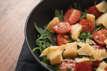 Fresh salad with cherry tomatoes, arugula and hard vintage cheese in black bowl on linen napkin