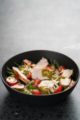Salad with chicken, tomatoes and arugula in black bowl on concrete background with copy space