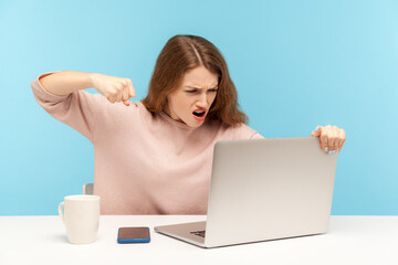 Enraged furious woman in casual clothes roaring madly and punching laptop screen, threatening with...