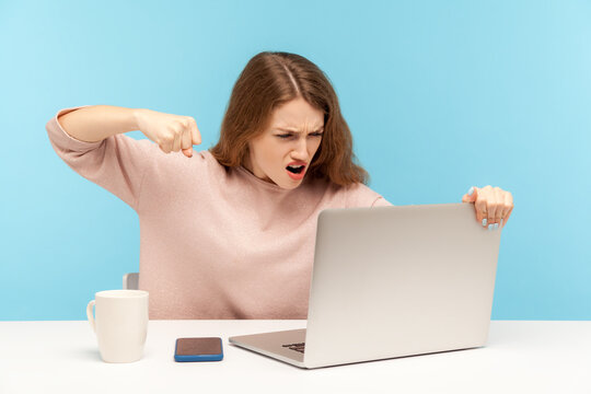 Enraged furious woman in casual clothes roaring madly and punching laptop screen, threatening with fist while talking on video call, online conference at home office. indoor studio shot, isolated