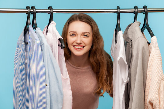 Lovely Positive Woman, Fashion Stylist Looking Out Of Clothes Hang On Shelf In Designer Store, Smiling Playfully And Hiding In Wardrobe With Trendy Outfit On Rack. Indoor Studio Shot, Isolated