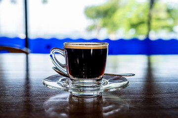 Transparent cup of coffee placed on wooden table near window, blurred green trees on background.