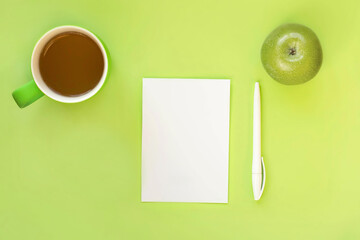 Top view of workspace. Blank sheet of paper, pen, apple and cup of tea on bright green background....